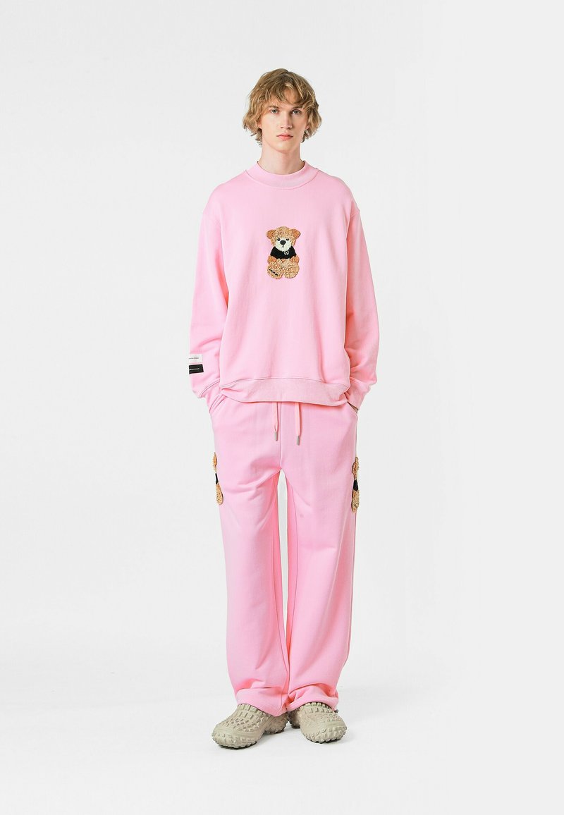Sudadera rosa con un diseño de oso, combinada con pantalones rosas que presentan bolsillos laterales. La tela parece suave y casual, con un corte relajado.