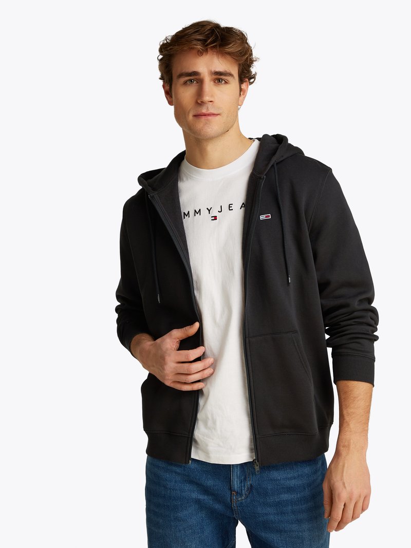 Haut de hoodie zippé gris foncé avec une poche avant et une capuche, porté par-dessus un t-shirt blanc avec un logo imprimé. Coupe décontractée, tissu en coton, poignets côtelés.