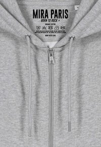 Felpa grigia con zip in cotone biologico, dotata di cappuccio regolabile, caratterizzata da una zip YKK e un design di etichetta nera a contrasto sul collo.