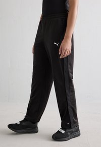 Calças de treino pretas com aberturas laterais de botões de estalo, cintura elástica e um logo branco da Puma na coxa esquerda. Usadas com ténis pretos.