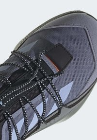 Shoe desportivo cinza com parte superior em malha, tecido texturizado, atacadores pretos e sola de borracha. Apresenta acentos azuis angulares e colar acolchoado.