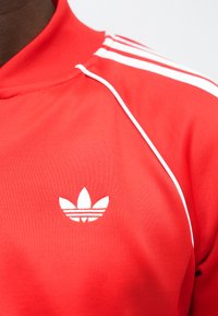 Casaco desportivo vermelho da Adidas com riscas brancas no ombro e logótipo trevo da Adidas no peito.