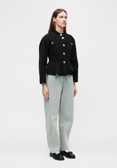 Γυναίκα που στέκεται φορώντας ένα μαύρο σακάκι με ασημένια κουμπιά, ανοιχτό γαλάζιο παντελόνι φαρδύ στο πόδι και μαύρα μοκασίνια, μπροστά σε απλό φόντο.