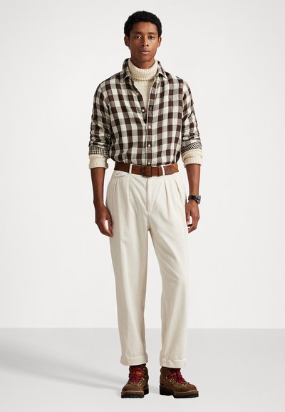 Camisa a cuadros en marrón y crema, cuello alto crema, pantalones de pana anchos en beige, cinturón marrón y botas de cordones marrones con acentos de colores.