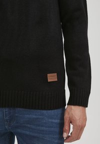 Pull en maille noir avec des poignets et un ourlet côtelés. Présente une étiquette marron en bas à gauche, indiquant la marque "11 PROJECT."