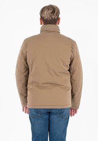 Uomo con capelli corti castano chiaro, che indossa una giacca marrone e jeans blu, in piedi con le braccia lungo i fianchi, rivolto di spalle.