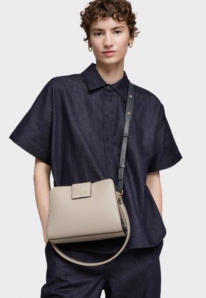 Mujer con camisa de manga corta de mezclilla oscura abotonada y pantalones, llevando un bolso Tous beige con correa negra cruzada sobre el hombro.