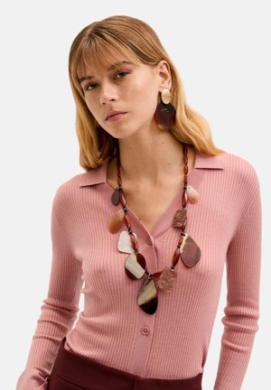 Perlekæde med store, farverige, ovale stykker i nuancer af brunt og creme, båret med en lyserød ribbet cardigan.