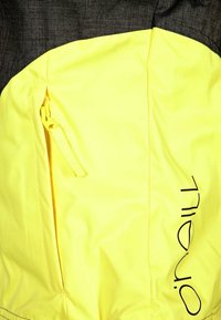 Giacca impermeabile gialla con una spalla texturizzata grigia scura, una tasca laterale con zip e un logo nero prominente sul davanti.