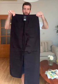 Black pants made of fabric, featuring a straight-leg design. A brand label is visible at the waistband. Background includes a sofa and flowers.