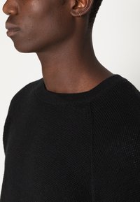 Suéter negro de punto con cuello redondo y patrón texturizado, mostrando un primer plano del área del hombro y el cuello. Se puede ver un pequeño pendiente dorado.