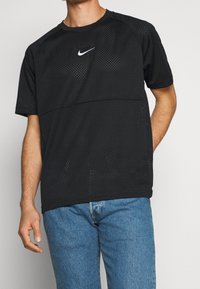 Svart perforerad träningsskjorta med korta ärmar och rundad halsringning, med en vit Nike-logga på bröstet. Matchad med blå jeans.