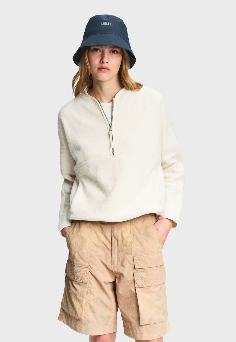 Woman wearing white fleece half-zip pullover, beige cargo shorts, and a navy bucket hat with "AIGLE" logo on a plain light background.