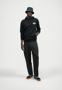 Hanorac negru cu buzunar frontal și grafică verde, pantaloni largi gri, pantofi negri și albi și o pălărie gri tip bucket. Fundal neutru.