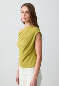 Haut asymétrique à une épaule en tissu vert clair, doté d'une texture drapée, de manches courtes et d'un design ajusté. Associe avec un pantalon blanc.