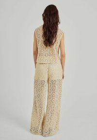 Beige háčkovaný outfit složený z bezramínkového topu a kalhot s širokými nohavicemi, na kterých jsou geometrické vzory. Tkanina má otevřenou, krajkovou strukturu.