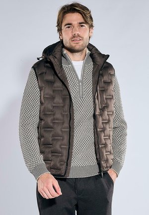 Mann trägt eine braune gesteppte Weste über einem gemusterten grauen und weißen Reißverschluss-Pullover, mit dunkler Hose, vor einem hellgrauen Hintergrund.