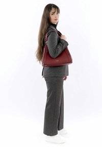 Rote Ledertasche mit glattem Material, runder Form und silbernen Hardware-Akzenten. Modell trägt einen grauen Anzug und weiße Sneakers.