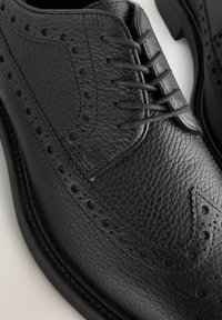 Chaussures habillées en cuir noir avec une finition texturée, présentant un design à bouts fleuris, des perforations décoratives et six œillets pour les lacets.