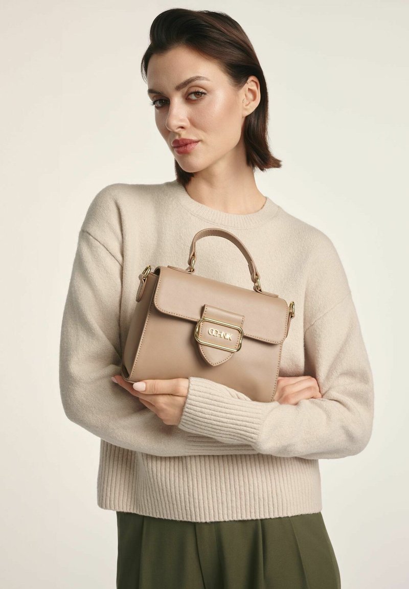 Skórzana torebka w kolorze brązowym z złotymi elementami, o strukturalnym wzorze, z górnym uchwytem i zamknięciem na klapę. Noszona z beżowym swetrem i zielonymi spodniami.