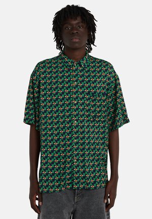 Person trägt ein kurzärmeliges Hemd mit grünem, marineblauem und orangem Muster, locker geschnitten, und dunkelgraue Hosen.