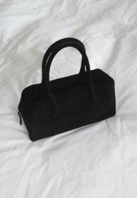 Melnas zamšādas rokassomiņa ar divām rokturiem un rāvējslēdzēju. Piedāvā kvadrātveida formu un gludu tekstūru. Minimālisma dizains bez acīmredzamām rakstu zīmēm.