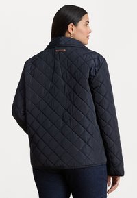 Quiltet marineblå jakke med krage og et brunt merke bak, designet med diamantmønster og lett tekstur.