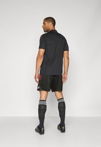 Zwarte polo met rode accenten, zwarte shorts, zwarte sokken met witte strepen en zwarte voetbalschoenen. Het ontwerp is sportief en aansluitend.