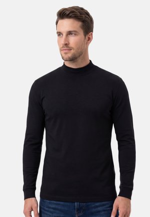 Jonge man met kort bruin haar, gekleed in een zwart shirt met een col en lange mouwen en een blauwe spijkerbroek, kijkt iets naar links tegen een effen achtergrond.