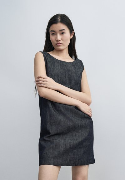 Jeune femme aux longs cheveux foncés portant une robe en jean foncé sans manches, debout les bras croisés devant un fond gris uni.