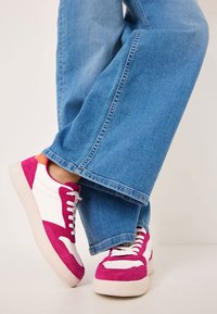 Sneakers in suede rosa e pelle bianca con suola spessa, dotati di lacci rosa e dettagli arancioni, abbinati a jeans a gamba larga di colore azzurro chiaro.