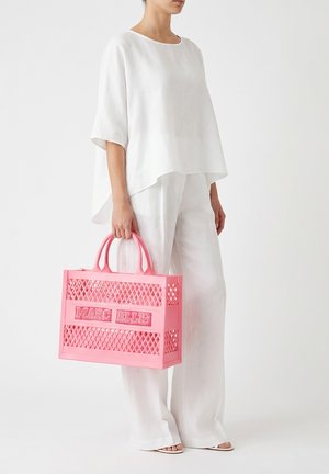 Personne vêtue d'un haut blanc ample et d'un pantalon tenant un grand sac cabas rose Marc Ellis avec un motif en treillis et des poignées.