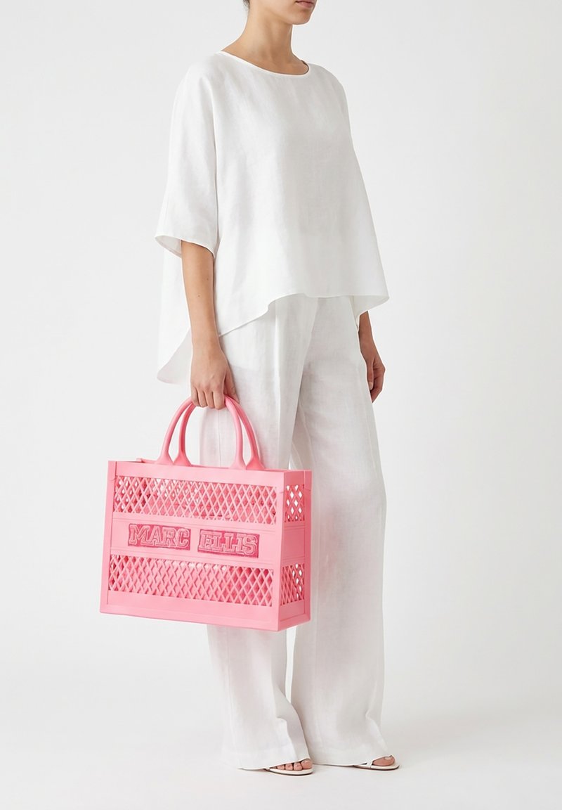 Personne vêtue d'un haut blanc ample et d'un pantalon tenant un grand sac cabas rose Marc Ellis avec un motif en treillis et des poignées.