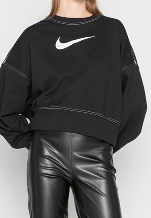 Osoba nosící černou mikinu Nike s bílým prošíváním a černé kožené kalhoty, stojící s jednou rukou v bok.