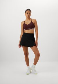 Burgundi sportmelltartó fehér Nike logóval, fekete, testhez simuló szoknyával párosítva. A modellen fehér sportcipők vannak, lime zöld részletekkel.
