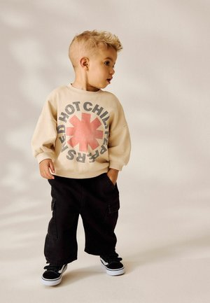 Bambino con capelli ricci corti, che indossa una felpa beige dei Red Hot Chili Peppers, pantaloni cargo neri e scarpe nere e bianche.