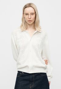 Νεαρή γυναίκα με ξανθά μαλλιά που φοράει γυαλιά με διάφανο πλαίσιο, άσπρο πουλόβερ με κουμπιά και διακριτικά σχέδια και σκούρο μπλε τζιν, σε λευκό φόντο.