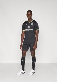 Juoda trumpomis rankovėmis futbolo marškinėlių su žaliais ir geltonais akcentais, liūto logotipu ir rėmėjo užrašu. Derinama su juodais šortais ir kojinėmis.