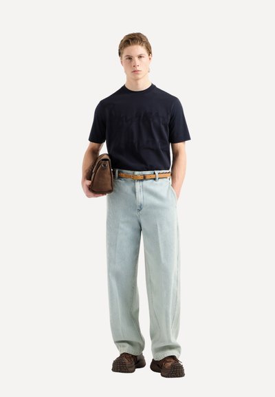 T-shirt navy à manches courtes, pantalon plissé léger bleu relaxé, ceinture marron, sac marron et chaussures texturées, se tenant devant un fond uni.