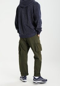 Mörkblå fleecetröja med huva, i kombination med olivgröna cargobyxor med sidofickor, och mörkblå/vita sportskor.