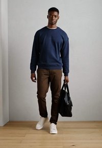Navy sweatshirt, brown pants, and white sneakers. Model holds a black leather bag. Simple design and neutral color palette.