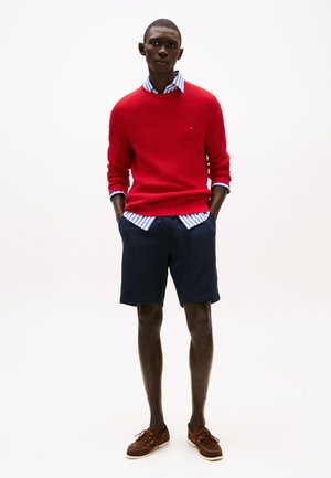 Red knitted sweater over a blue and white striped shirt, paired with navy shorts and brown boat shoes. Simple design and contrasting textures.