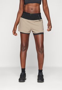 Käbe tan lühikesed püksid musta vöökoha ja aktsentidega, millel on tekstuuritud muster ja pael. Kombineeritud mustade spordijalatsite ja sokkidega.