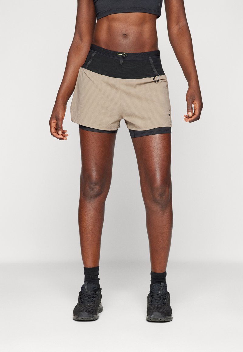 Käbe tan lühikesed püksid musta vöökoha ja aktsentidega, millel on tekstuuritud muster ja pael. Kombineeritud mustade spordijalatsite ja sokkidega.