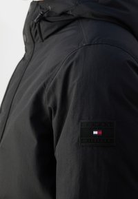 Musta hupparillinen takki, jossa on sileä tekstuuri ja Tommy Hilfigerin merkki hihaisessa suorakaiteen muotoisella designilla ja logon väreillä.