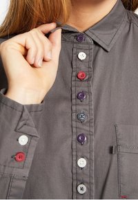 Grå skjorte med knapper og spids krave, kontrasterende knapper i rød, hvid, lilla og blå, med en brystlomme på venstre side.