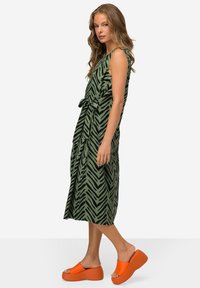 Grünes Kleid mit schwarzem Zickzackmuster, ärmelloses Design, mit Bindegürtel in der Taille, kombiniert mit orangefarbenen Plateauslippern. Elegante Midi-Länge.