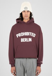 Hombre con un hoodie burdeos que tiene el texto "PROHIBIDO BERLÍN", pantalones grises con pliegues, un gorro de punto color crema y anillos de plata en los dedos.