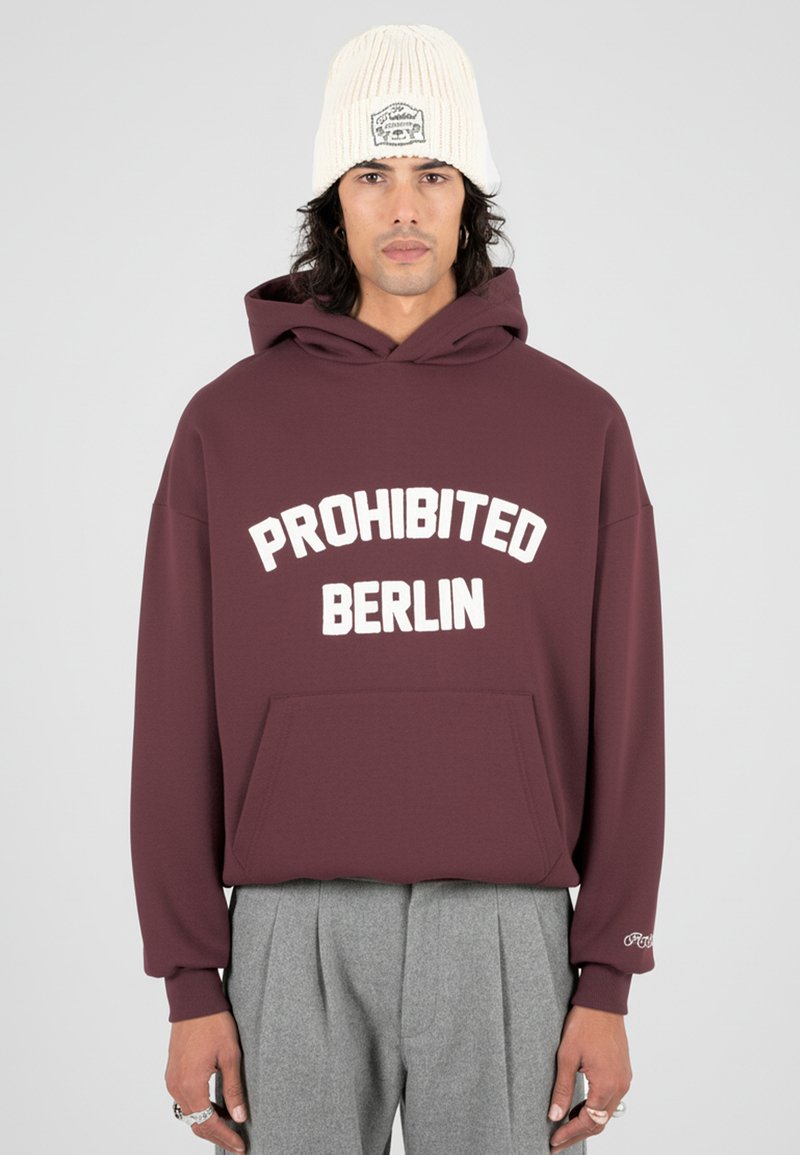 Hombre con un hoodie burdeos que tiene el texto "PROHIBIDO BERLÍN", pantalones grises con pliegues, un gorro de punto color crema y anillos de plata en los dedos.