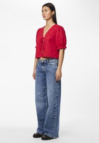 Cropped top rosso con scollatura a V, maniche a sbuffo e chiusura con lacci, abbinato a jeans a vita alta con gamba larga di un blu sbiadito e calzature nere.
